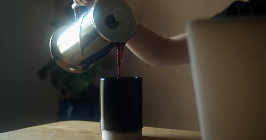 A woman sitting in front of a laptop pours coffee from a coffee pot into a mug before lifting it to take a drink