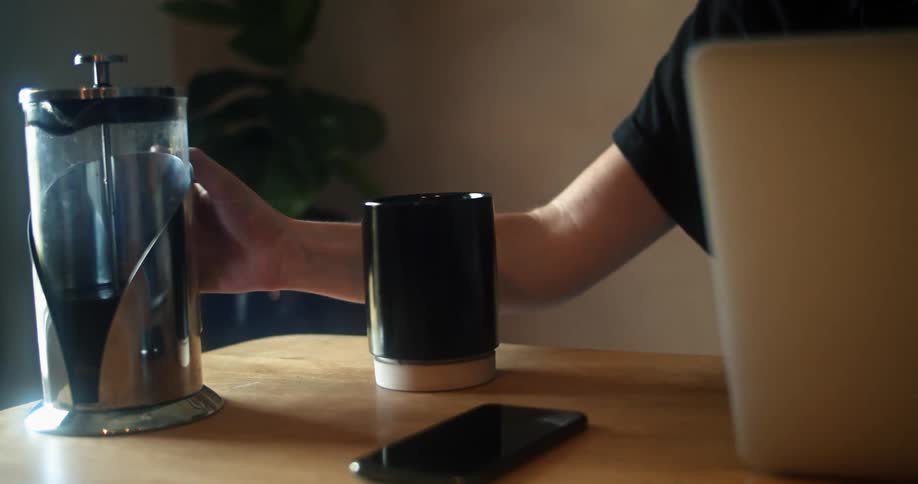 A woman sitting at home in front of her laptop pours herself a cup of coffee and takes a drink