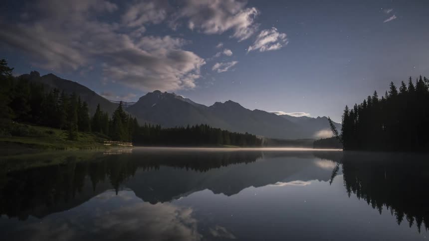 Still waters of lake bordered by forest and mountains reflects surrounding including sky