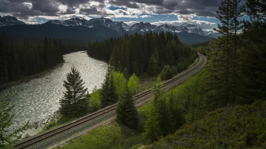 Train follows train tracks away from mountains past forest and paralleling river
