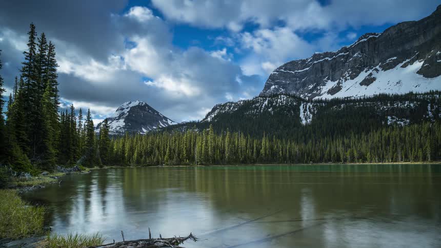 The rippling surface of a lake at the base of a mountain reflects the surrounding forest and the clouds passing overhead as the day progresses