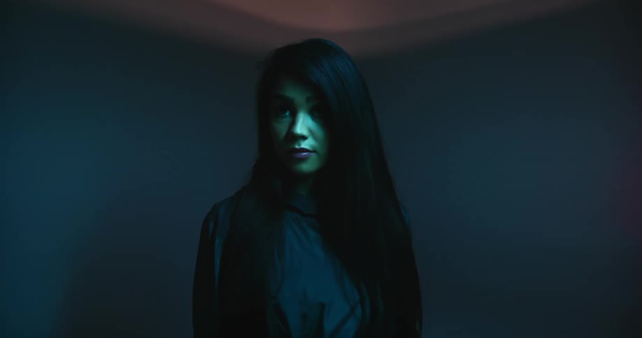 A teenage girl with long black hair stands in the middle of a room not moving or blinking as a red light circles around her