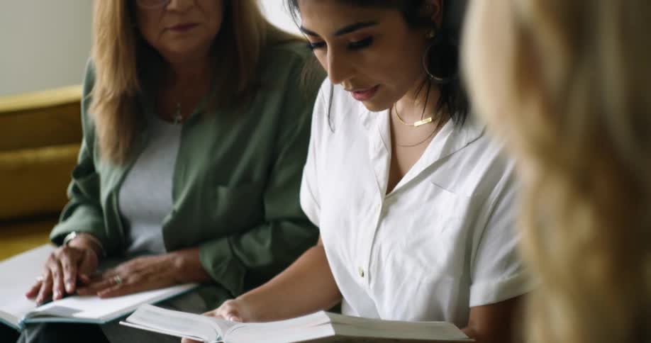 A young woman reads from a large textbook as two women seated beside her ready to take notes