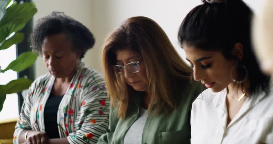 Three women sitting on a couch and praying with a window in the background