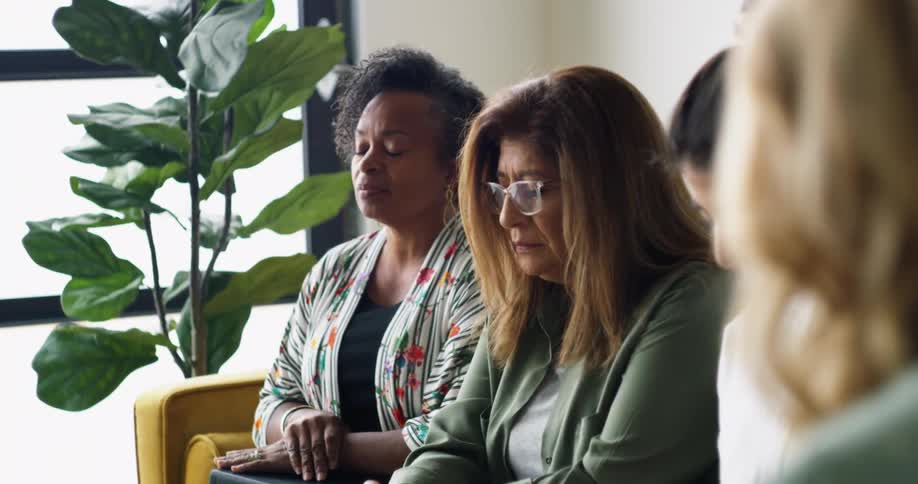 Four women of different ethnicities sit on a yellow couch together and pray