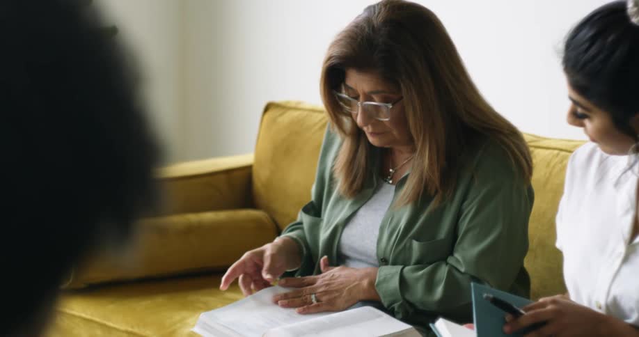 A woman looks on as another turns the pages of a book in her lap