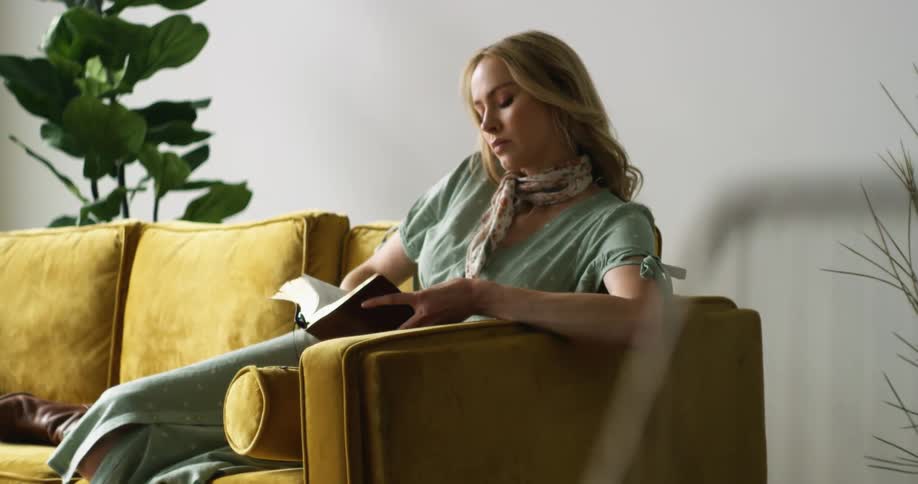 Blonde woman sitting in a yellow sofa reads a book in the living room at home