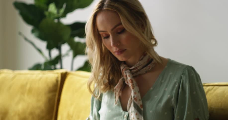A blonde woman sitting on a sofa looks up and turns her head toward the light of a window