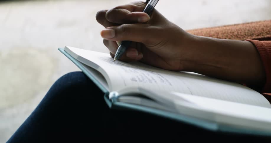 A person sits in a chair indoors while writing in a hardcover notebook