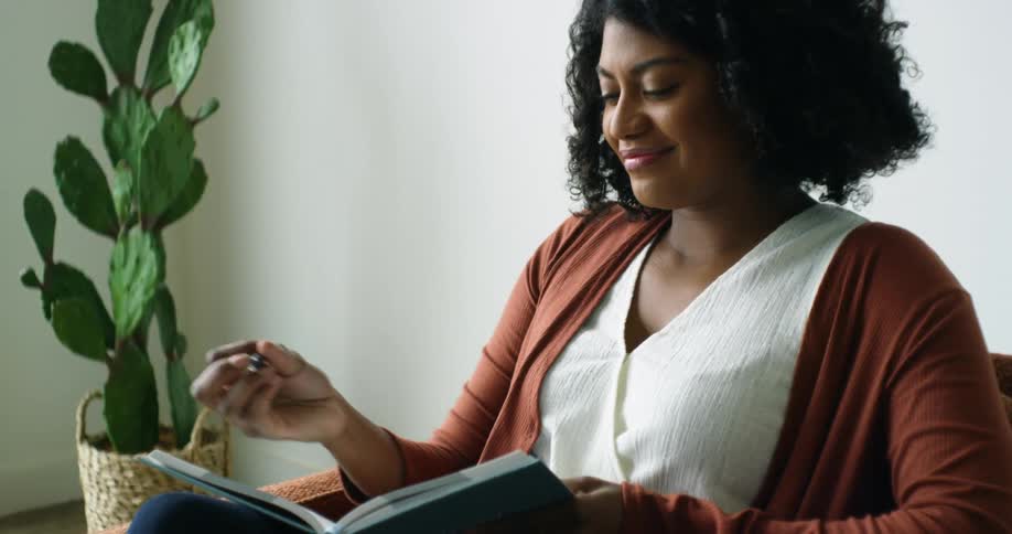 A woman sitting in a chair smiles as she looks down and begins to write in a journal