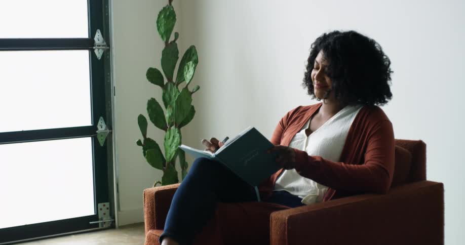 Black woman with curly hair sitting in a red armchair writes in a notebook smilingly