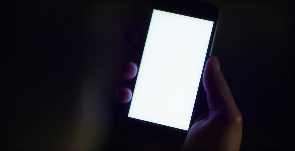 Person holds mobile phone with brightly lit blank screen in dark area