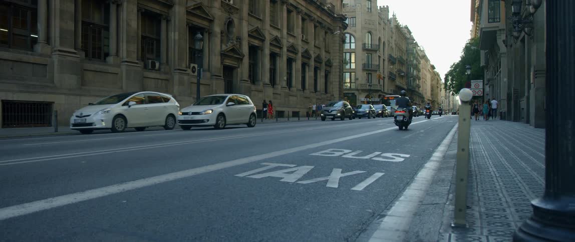 People walk down a city sidewalk as cars and motorcycles drive past