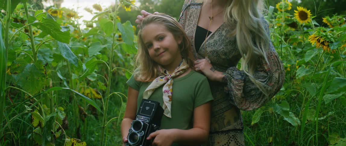 A girl holding a camera with a woman fixing her hair with flowers trees and the sky in the background