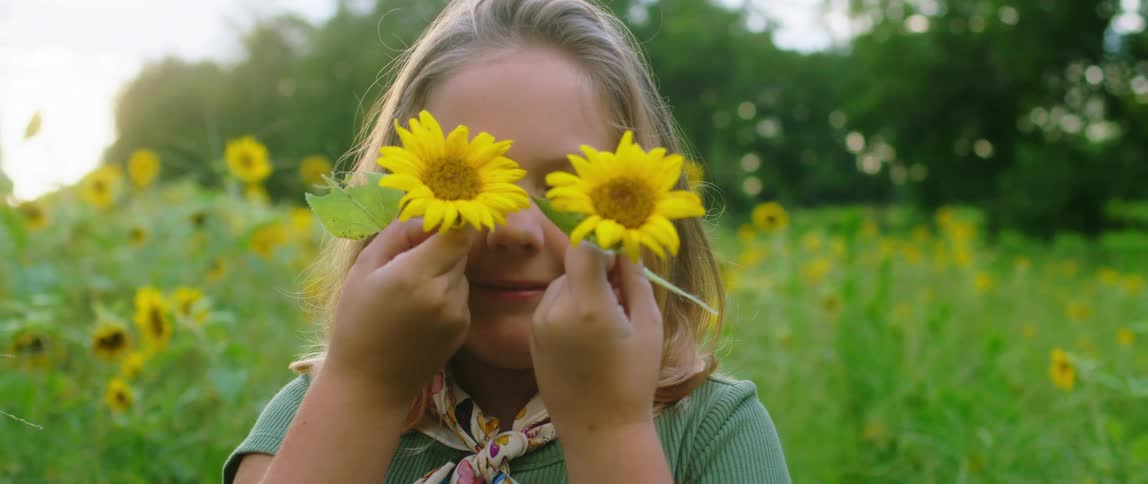 A girl standing in a grassy field holding two flowers up to her face with trees in the background