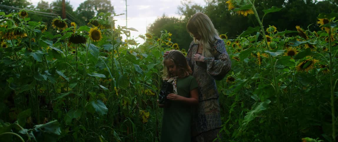 Mother and daughter stand in field among sunflowers as daughter uses camera