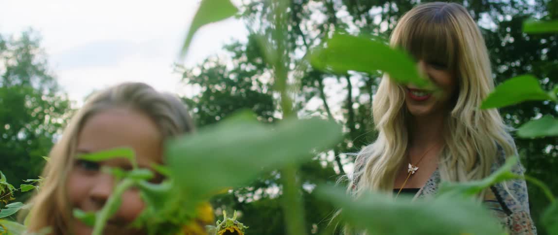 A woman and a girl standing and surrounded by flowers with trees and the sky in the background