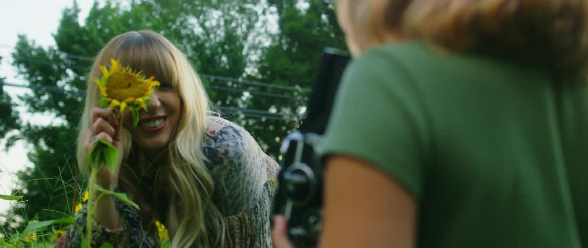 A mother holds a sunflower over her face and looks at her daughter who is holding a camera