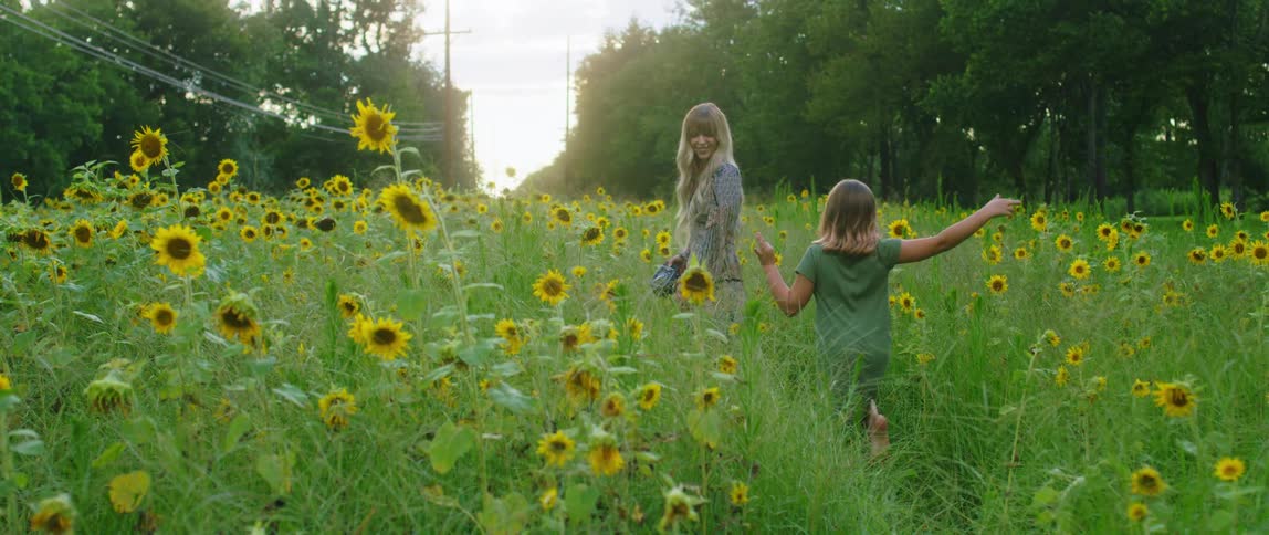 A mother carries a camera as her and her daughter walk through a field of sunflowers