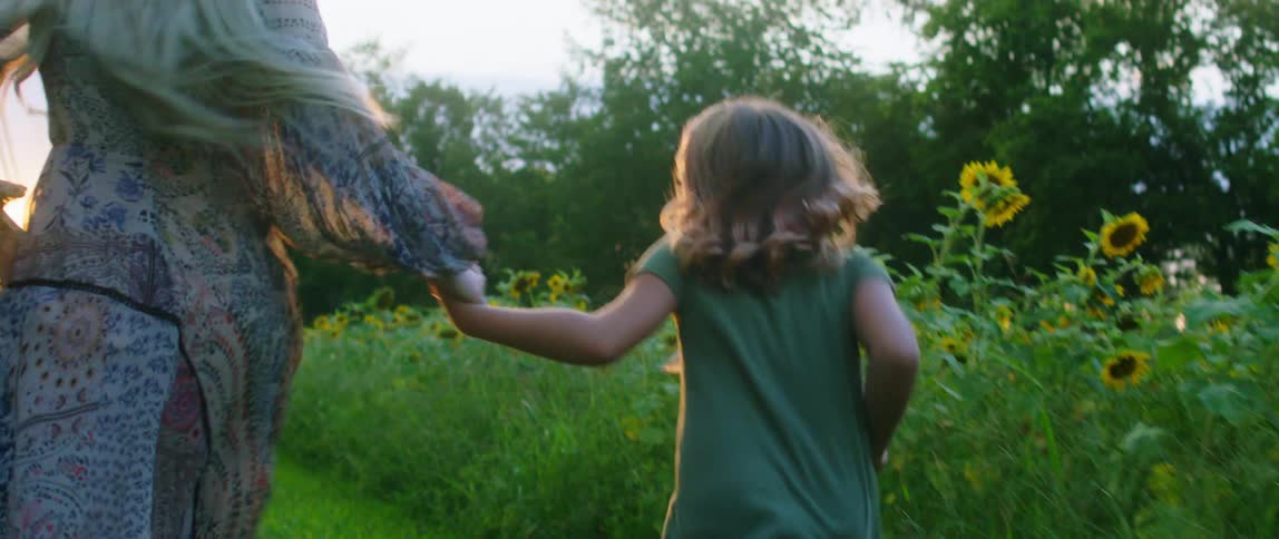 A mother and daughter run in the grass next to sunflowers while holding hands