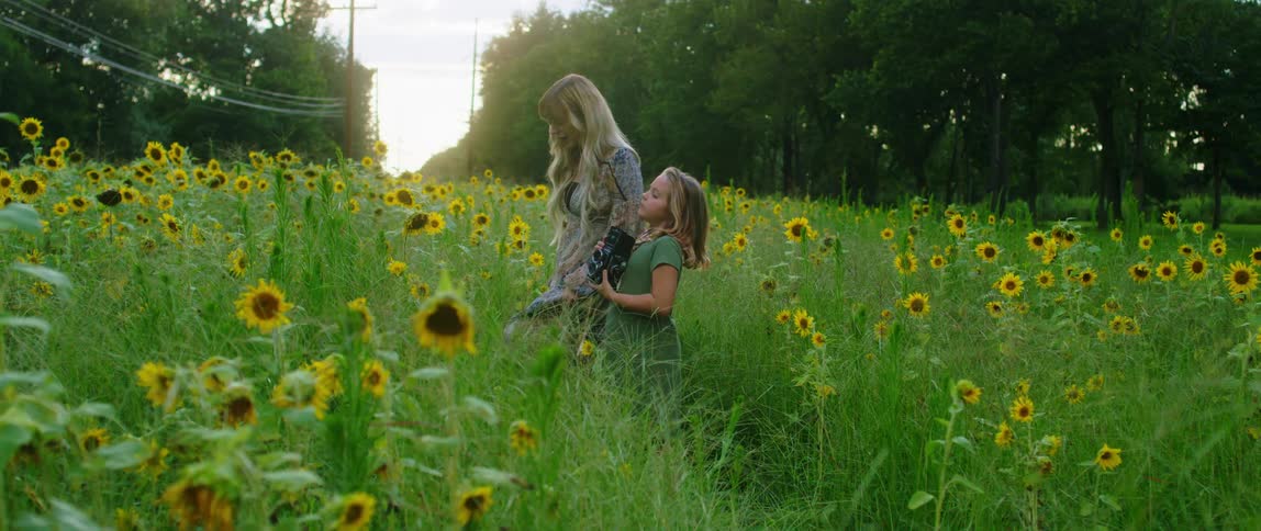 A white woman and white child stand in a grass field full of sunflowers