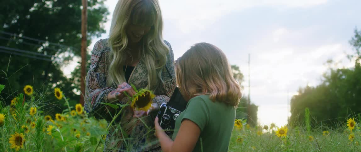 A girl holding a camera standing in a grassy field with flowers next to a woman