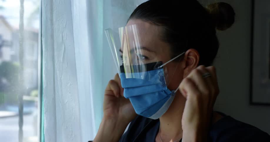 Female nurse wearing a scrub looks out the windows and puts on a blue mask