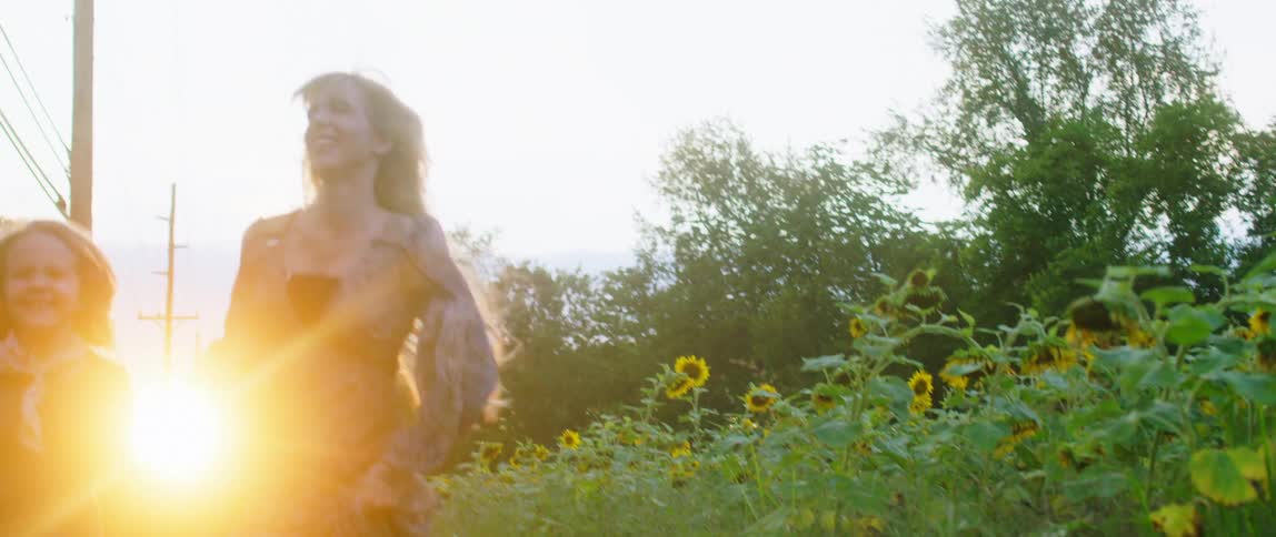 A mother and daughter holding hands and run barefoot through the grass next to sunflowers