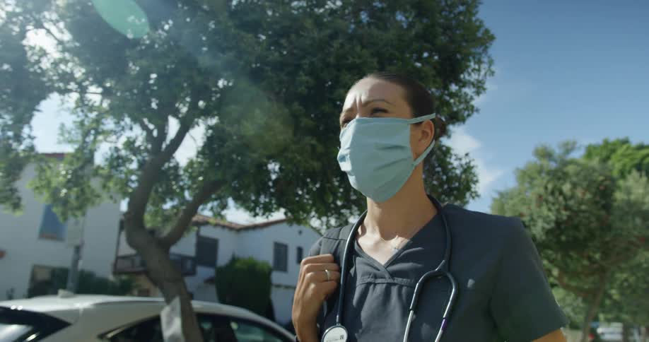 A woman with a mask a stethoscope and scrubs walking with houses and trees in the background
