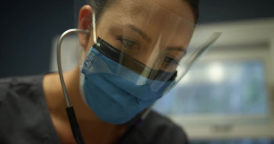 Nurse wearing scrubs with face mask and face shield listens through stethoscope