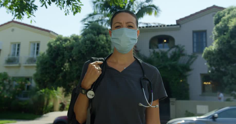 A woman with a mask stethoscope and scrubs standing with houses and trees in the background