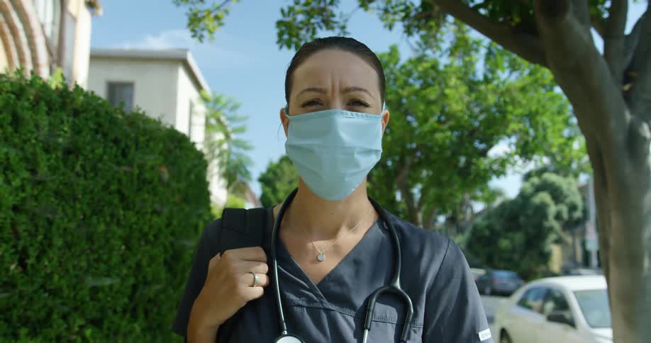 A nurse standing outdoors in a city neighborhood with a backpack over one shoulder and wearing a medical mask squints into the camera