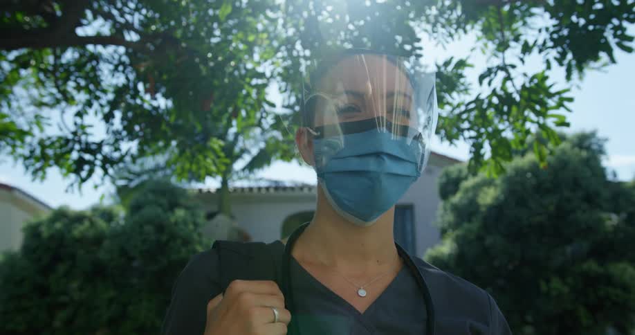 A white woman stands outside a house wearing a mask and a face shield