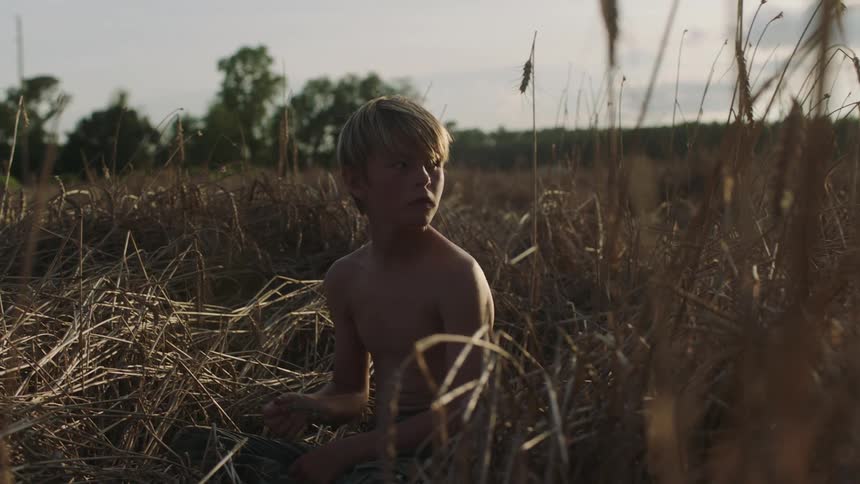 A young boy sitting with no shirt on in grass and hay