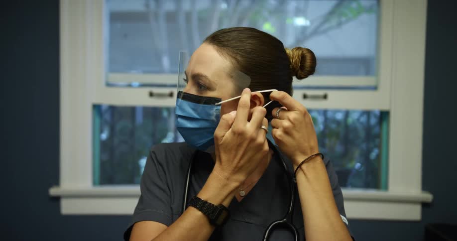 Woman in scrubs near window with stethoscope around her neck adjusts face mask with face shield
