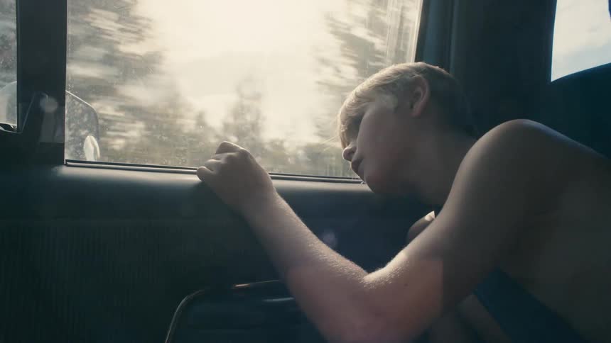 A shirtless boy rests his head on the door as he rides in the back seat of a vehicle