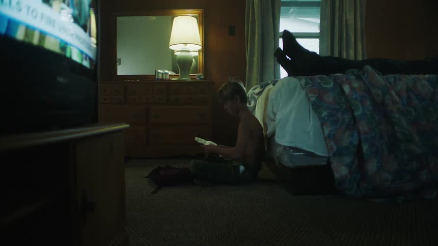 A young boy sits at the foot of a bed reading a paper while his parent lies in bed watching television