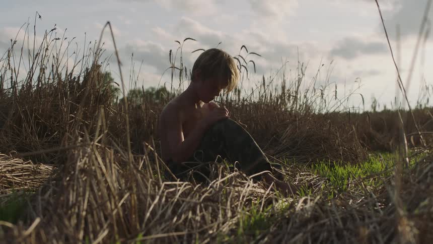 A boy with jeans sitting on grass with the sky and clouds in the background