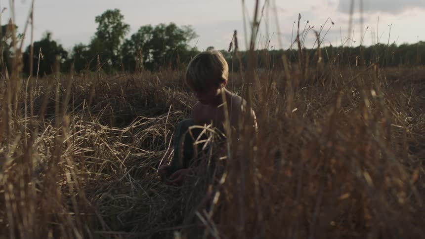 Bare chested boy sits on ground in field of dried grass under a cloudy sky