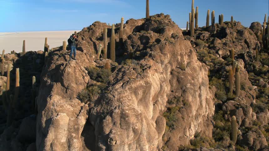 A photographer takes a photograph while standing atop a large rock formation in the desert on a sunny day