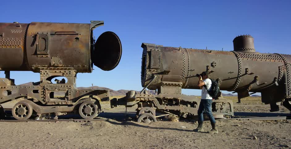 A photographer takes photographs of a derelict train in the desert on a sunny day