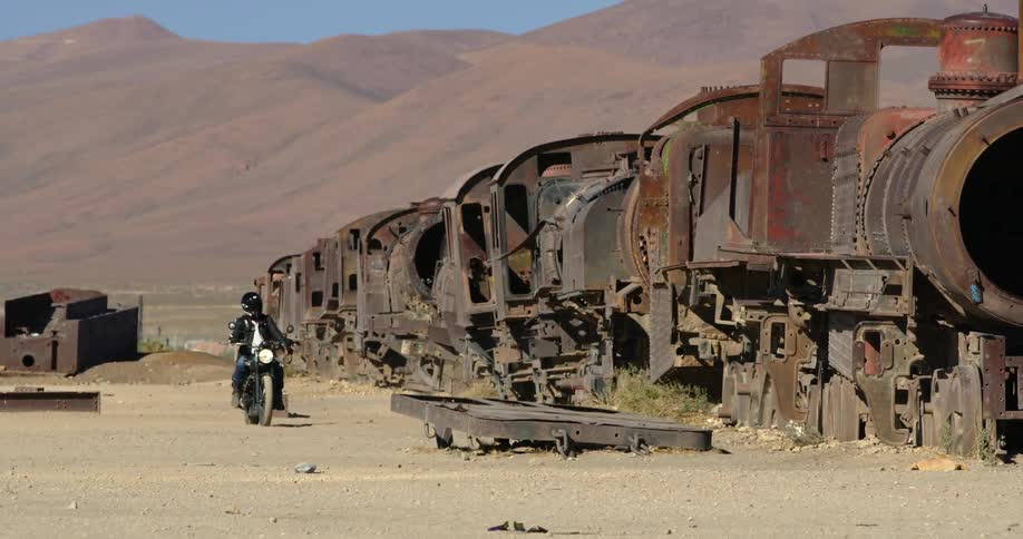 Man rides motorcycle across sand along length of abandoned train near mountainunder clear blue sky