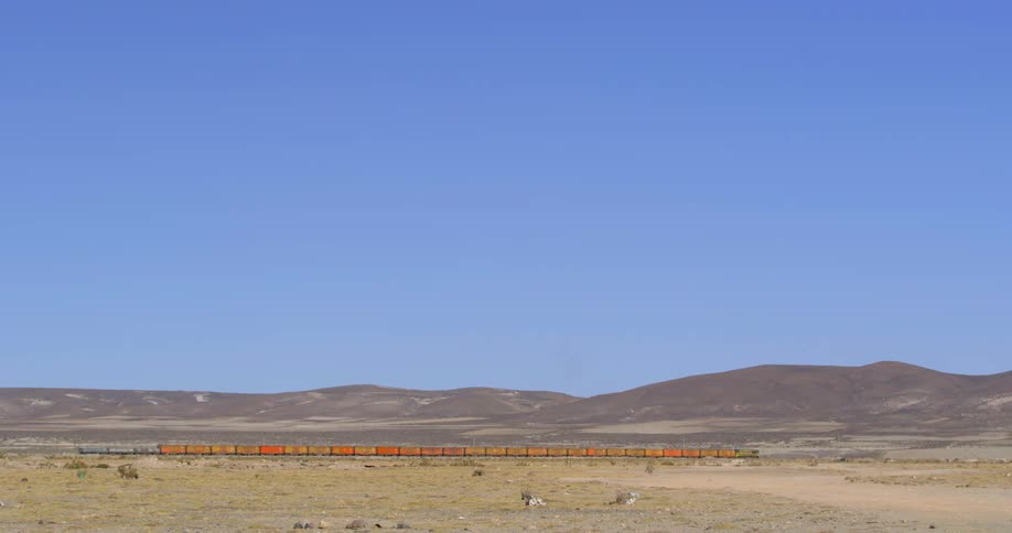 A red freight train passes by hills on one side and flat terrain on the other
