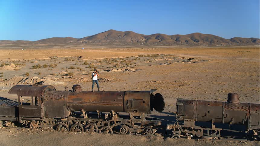 A photographer walking atop a derelict train in the desert stops to take a photograph before continuing onward