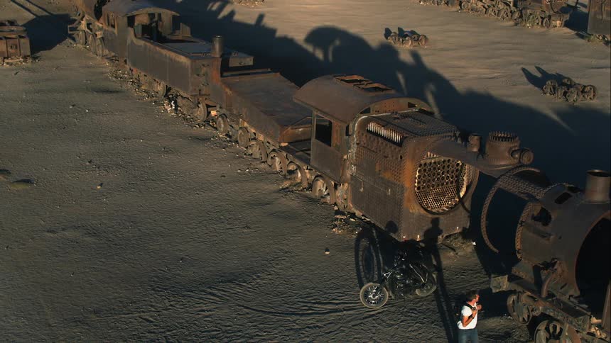 A man takes his jacket off and puts it on his motorcycle and walks to examine an old broken down train