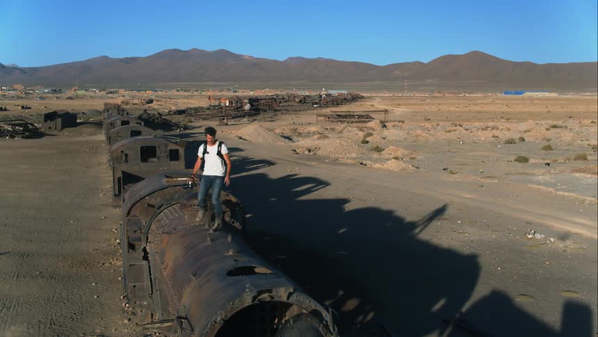 A photographer walks across the roof of a derelict train in a desert and raises his camera to take a photograph upon reaching the front