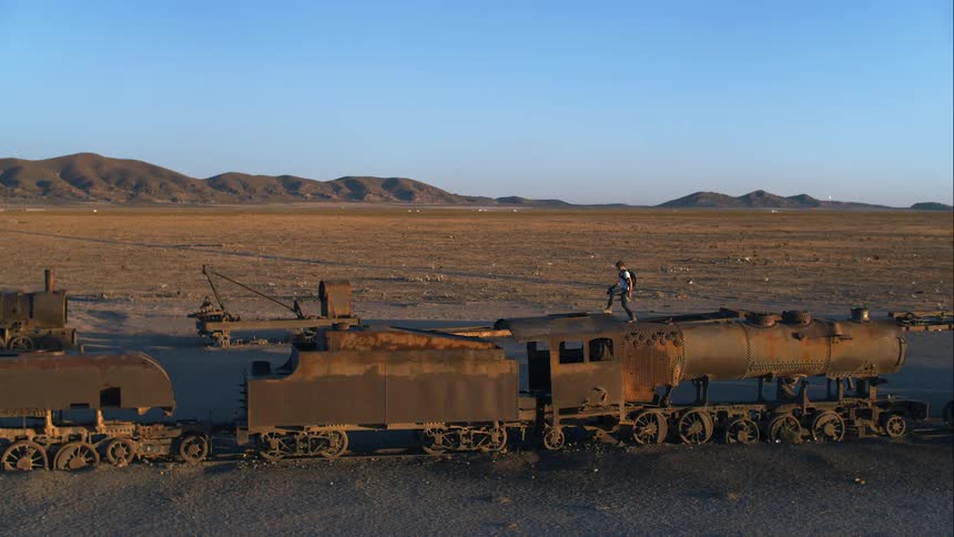 A photographer walks around a derelict train abandoned in the desert on a sunny day