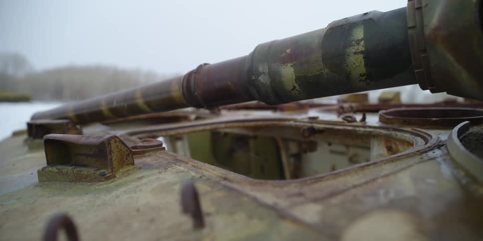 A rusted tank with an open top entrance sits in a field near a forest