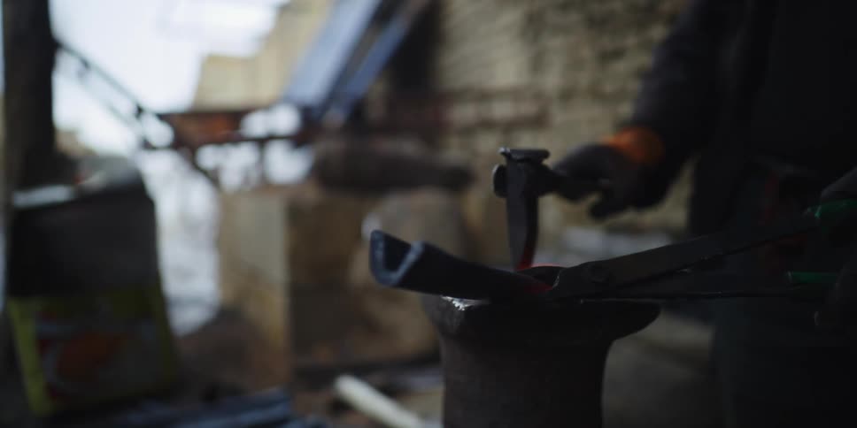 One person holds metal with tongs while another hammers it