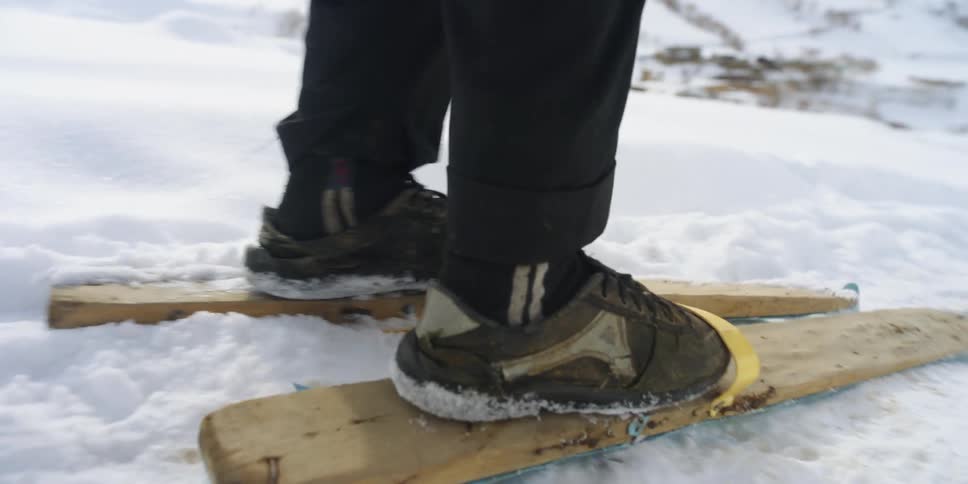 A person attempts to ski down a mountain slope atop a pair of ordinary wooden planks strapped to their sneakers